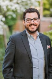 Smiling man with dark curly hair, beard, and glasses wearing a grey blazer outdoors.